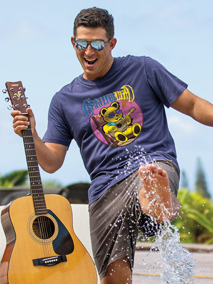 Man in a Grateful Dead blue t-shirt with a bear graphic holding a guitar, standing in water outdoors.
