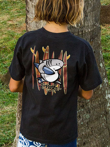 Child wearing a black t-shirt with a shark and surfboard design, standing next to a tree.