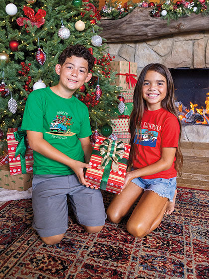 Two children in front of a Christmas tree with presents, wearing festive shirts.