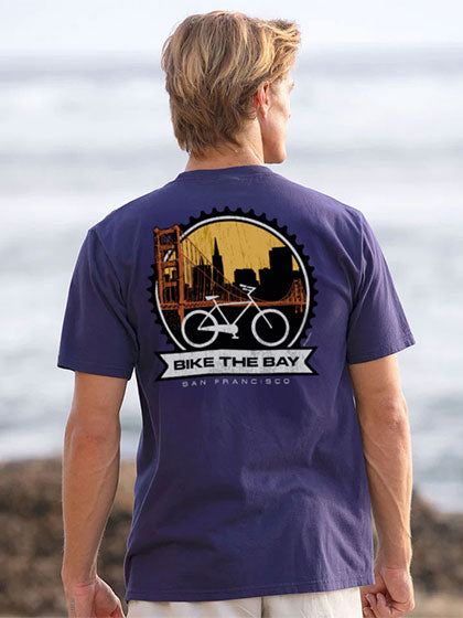 Person wearing a purple t-shirt with 'Bike the Bay' design by the ocean