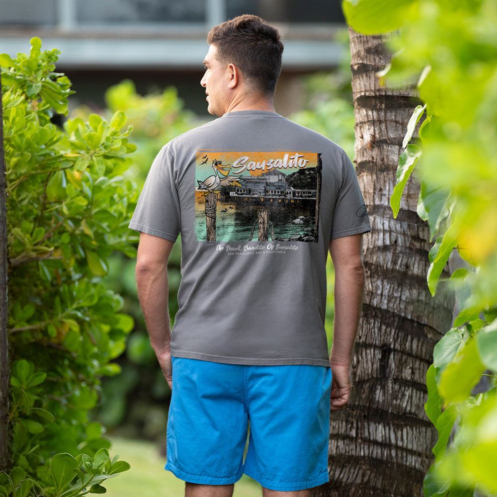 Man wearing a gray t-shirt with a colorful graphic on the back, standing outdoors with greenery around.
