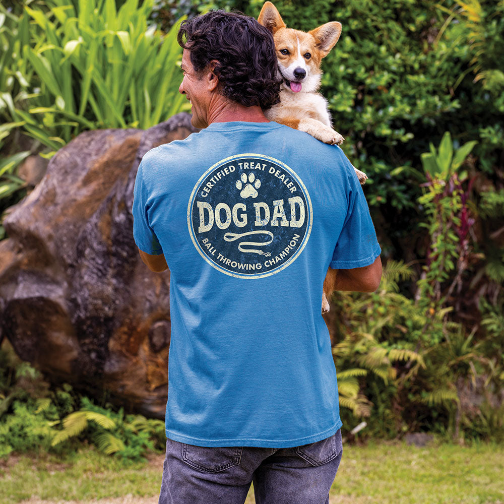 Man wearing a blue 'Dog Dad' t-shirt with a corgi on his shoulders, surrounded by greenery.