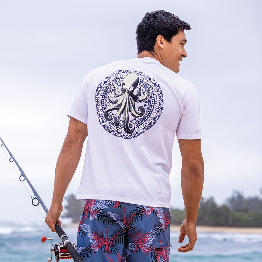 Man wearing a white t-shirt with an octopus design and blue patterned shorts, holding a fishing rod by the water.