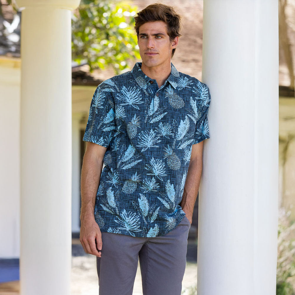 Man wearing a blue patterned shirt standing between white columns outdoors.