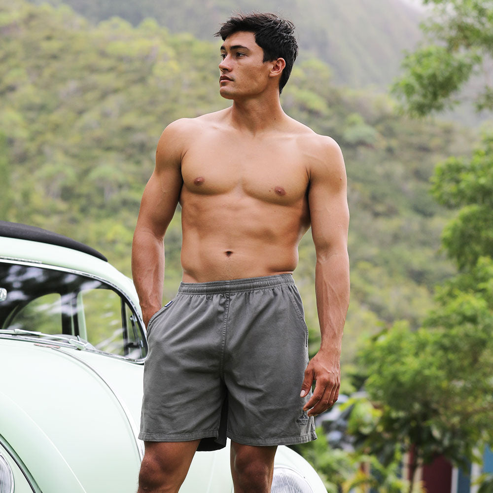 Man wearing light green swim shorts standing on a porch with a surfboard and chair in the background.