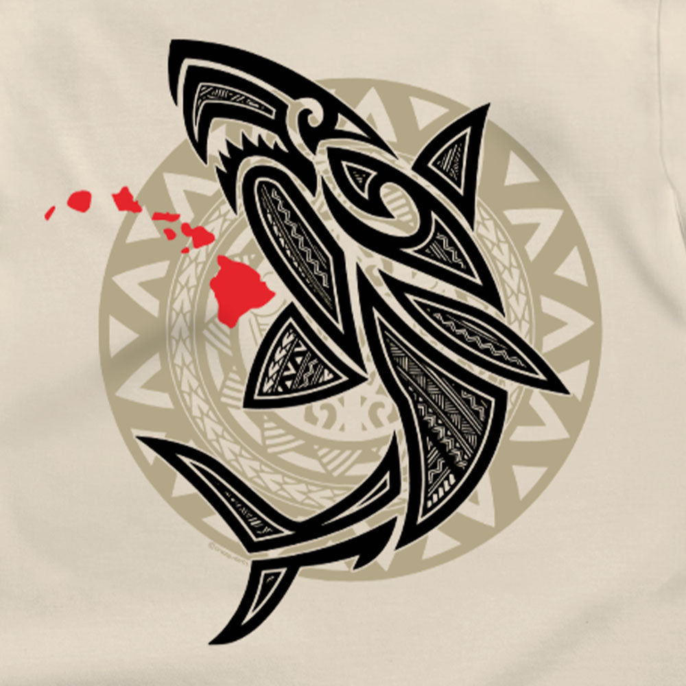 A close-up of a beige garment featuring a large Polynesian-inspired shark graphic in black, with red Hawaii silhouettes and a circular tribal pattern in the background.