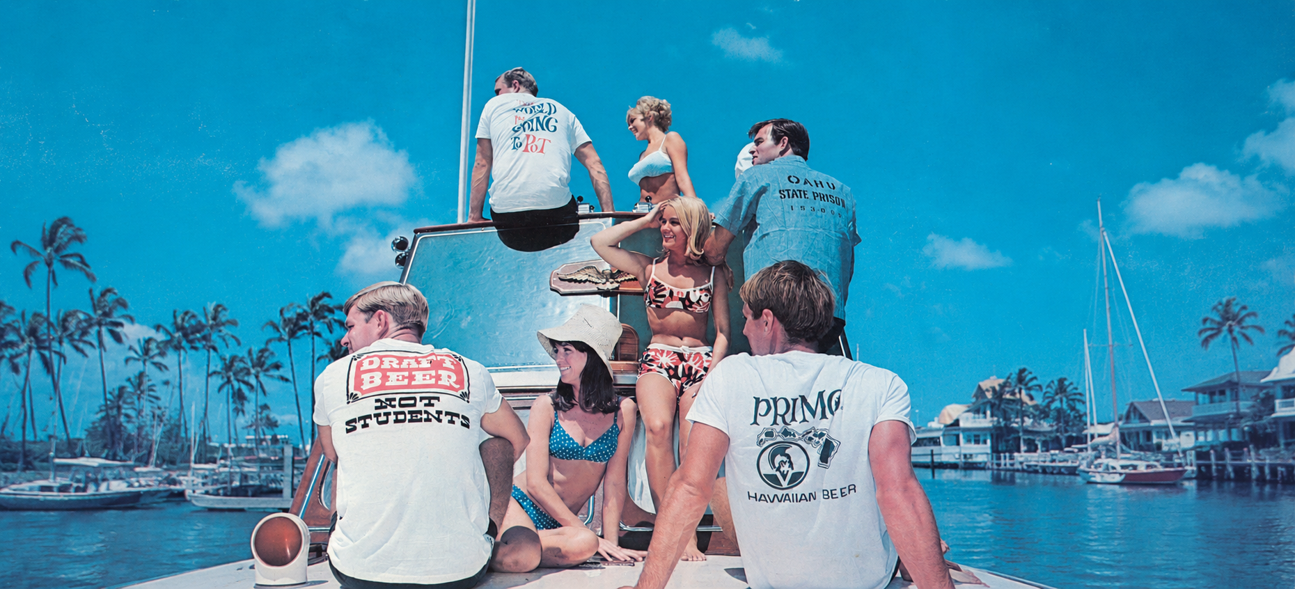A group of young men and women sitting and standing on the deck of a boat in a tropical harbor, wearing matching white t-shirts with various graphic designs and text.