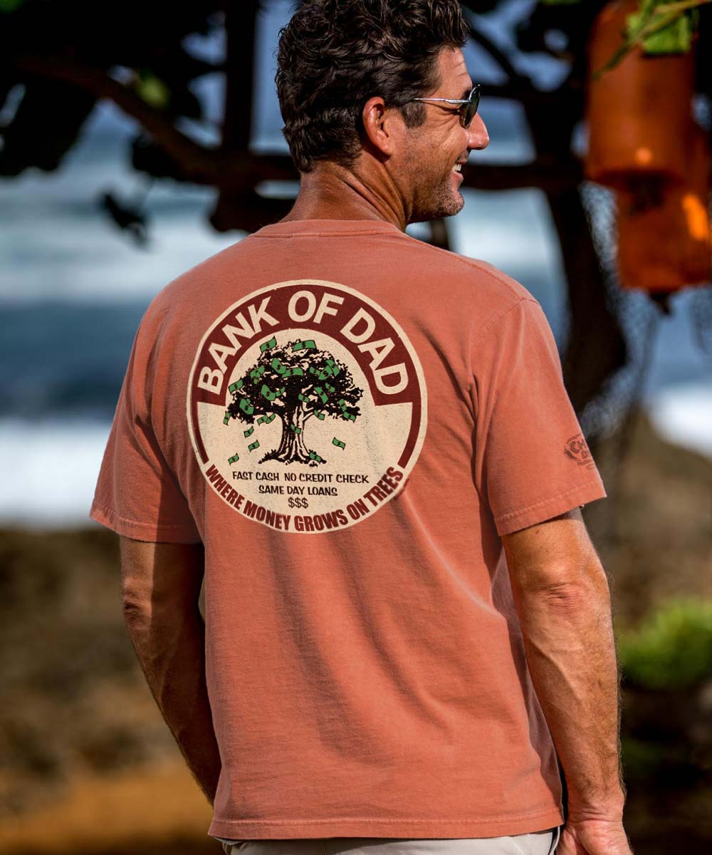 A man wearing a salmon short sleeve crewneck t-shirt with the text '_BANK OF Dad' printed on the back, featuring a tree illustration.