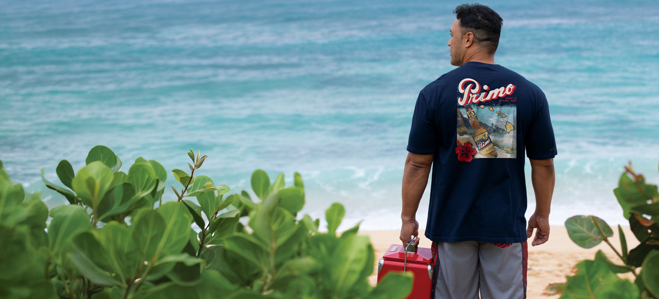 Man standing on a beach with a blue shirt featuring graphics and text, looking out at the ocean.